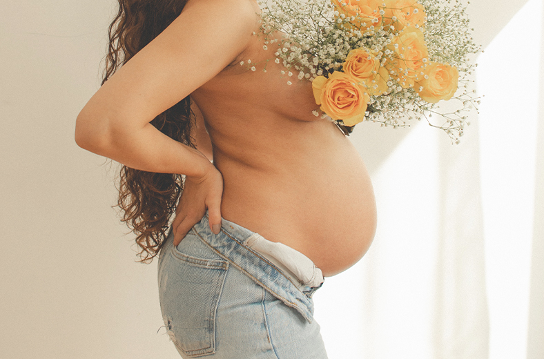 Cuerpo de perfil de una mujer embarazada con el torso desnudo y un ramo de rosas amarillas sobre el pecho